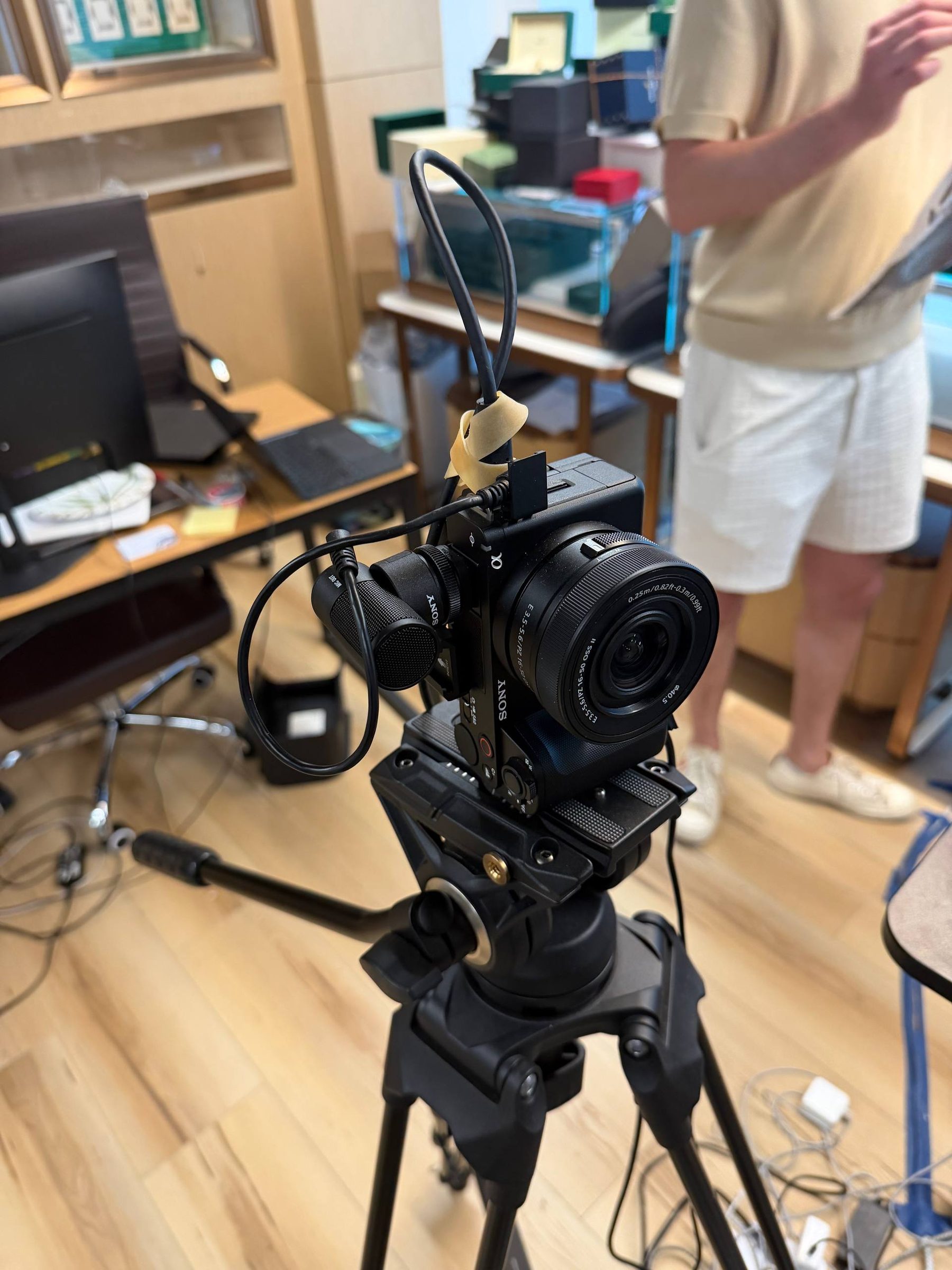 Video camera on a tripod in a watch showroom, crew member standing behind