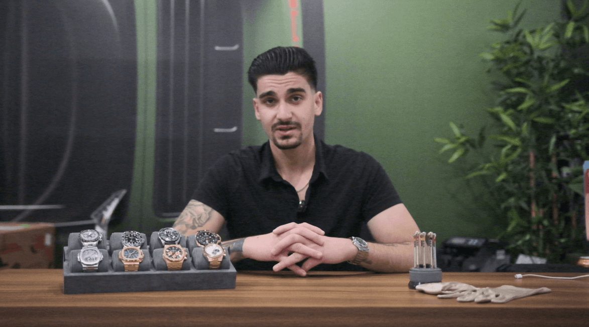 Before: presenter seated at a wooden desk against a green wall, a tray of watches in front of him