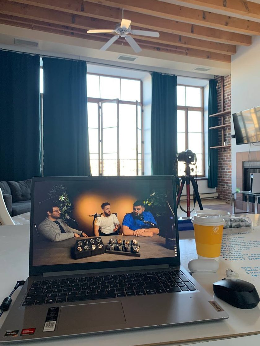 Laptop showing three presenters at a table with watches, camera on tripod behind